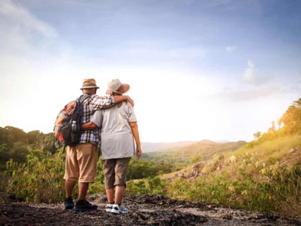 A pair of senior hikers put their arms around each other as they stare off into the sunset over rolling hells